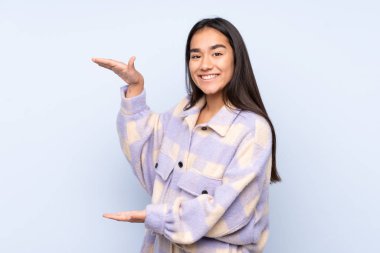 Mavi arka planda izole edilmiş genç Hintli bir kadın ilan vermek için kopyalama alanı tutuyor.