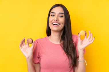 Genç esmer kadın izole edilmiş sarı arka planda elinde renkli Fransız kurabiyeleri tutuyor.