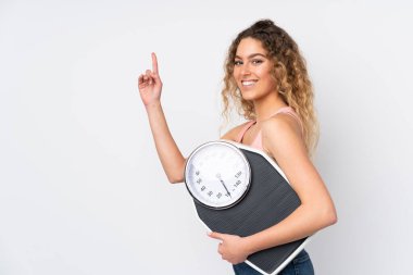 Beyaz arka planda izole edilmiş kıvırcık saçlı, tartı makinesi olan ve işaret eden genç sarışın kadın.