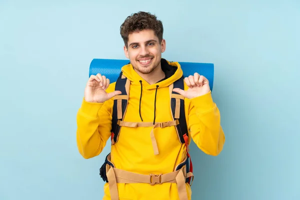 青の背景に誇りと自己満足感に隔離された大きなバックパックを持つ若い登山家の男 — ストック写真