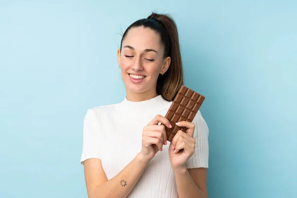 Young Brunette Woman Isolated Blue Background Taking Chocolate Tablet Happy — Stock Photo, Image