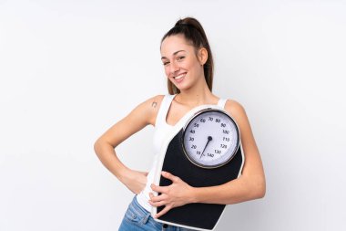 Genç esmer kadın izole edilmiş beyaz arka planda, kolları kalçasında ve tartı makinesini tutuyor.