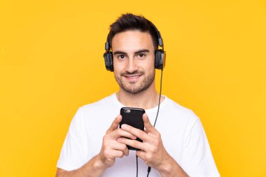 Sarı arka plandaki genç adam cep telefonuyla müzik dinliyor ve ön tarafa bakıyor.