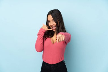 Mavi arka planda izole edilmiş genç melez bir kadın telefon hareketi yapıyor ve ön tarafı işaret ediyor.