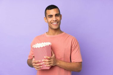 Genç Afrikalı Amerikalı bir adam mavi arka planda elinde koca bir kova patlamış mısır tutuyor.