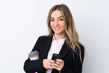 İzole edilmiş beyaz arka planda genç bir iş kadını elinde bir fincan kahve ve bir telefon tutuyor.