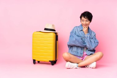 Genç gezgin Vietnamlı kadın bavuluyla yere oturmuş pembe arka planda yan tarafa bakıyor.