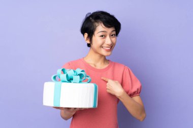 Kısa saçlı genç Vietnamlı bir kadın, izole edilmiş mor arka planda, yüzünde sürpriz bir ifadeyle büyük bir pasta tutuyor.