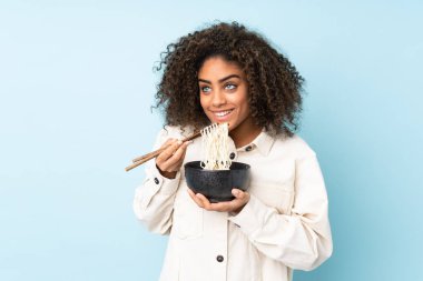 Mavi arka planda izole edilmiş genç Afrikalı Amerikalı kadın elinde yemek çubuklarıyla bir kase erişte tutuyor.