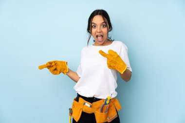 Mavi arka planda izole edilmiş genç elektrikçi kadın şaşırmış ve işaret ediyor.