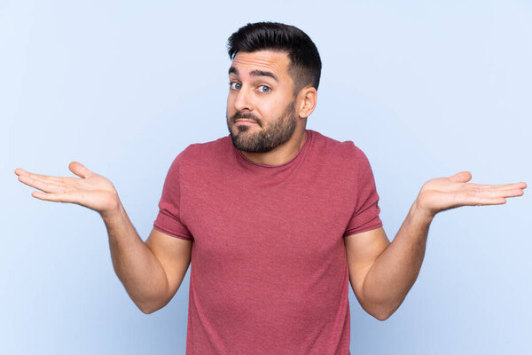 Young handsome man with beard over isolated blue background having doubts while raising hands