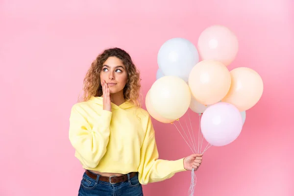Young blonde woman with curly hair catching many balloons isolated on pink background thinking an idea