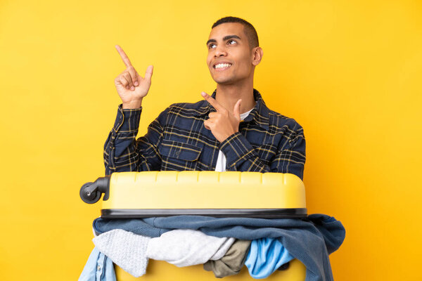 Traveler man with a suitcase full of clothes over isolated yellow background pointing with the index finger a great idea