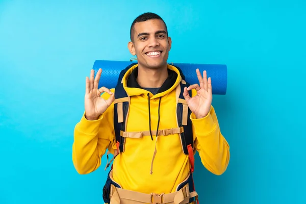 Молодой Альпинист Большим Рюкзаком Изолированном Синем Фоне Показывает Знак Пальцами — стоковое фото