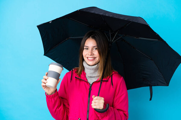 Young woman holding an umbrella and coffee to take away over isolated blue wall