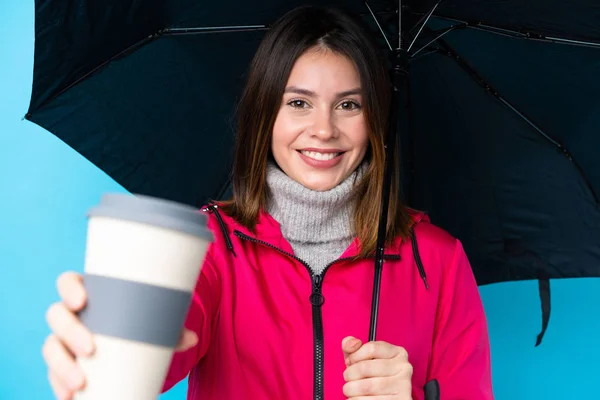 Young woman holding an umbrella and coffee to take away over isolated blue wall