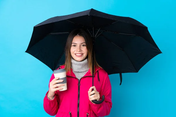 Young woman holding an umbrella over isolated blue wall smiling a lot