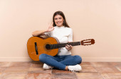 Mladá žena s kytarou sedící na podlaze a ukazující znamení ok s prsty