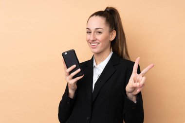 Cep telefonu kullanan genç iş kadını izole edilmiş arka planda gülümsüyor ve zafer işareti gösteriyor.
