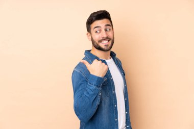Beyaz tenli yakışıklı adam bej arka planda izole edilmiş bir ürün sunmak için kenarı işaret ediyor.