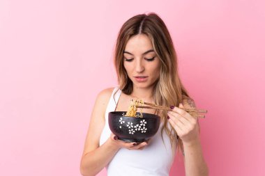 Soyutlanmış pembe arka planı olan genç bir kadın elinde yemek çubuklarıyla bir kase erişte tutuyor.