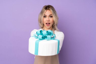 Genç pasta şefi mor arka planda büyük bir pasta tutuyor.