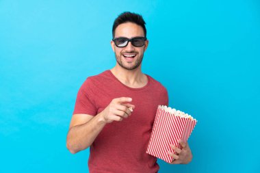 Üç boyutlu gözlüklü ve elinde koca bir kova patlamış mısırla, izole edilmiş mavi arka planda genç bir adam.