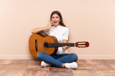 Gitarlı genç bir kadın yerde oturuyor ve yüzünde sürpriz bir ifade var.