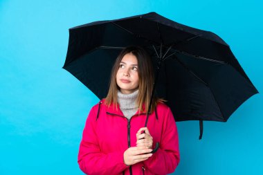 İzole edilmiş mavi duvarın üzerinde şemsiye tutan genç bir kadın omuzlarını kaldırırken şüpheyle hareket ediyor.
