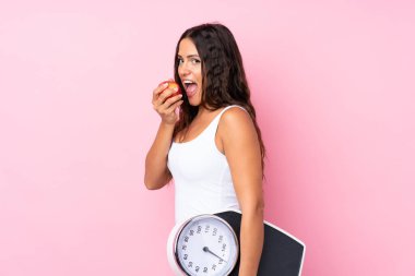 Soyutlanmış pembe arka plan, tartı makinesi ve elması olan genç bir kadın.