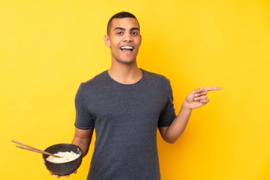 İzole edilmiş sarı arka planda yaşayan genç Afrikalı Amerikalı bir adam elinde çubuklarla bir kase erişte tutarken şaşırdı ve parmağıyla kenarı işaret etti.