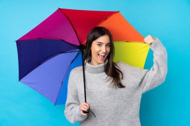 Genç esmer kadın mavi duvarın üzerinde şemsiye tutuyor güçlü bir jest yapıyor.