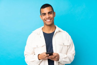 Genç Afro-Amerikalı bir adam mavi arka planda cep telefonuyla mesaj gönderiyor.