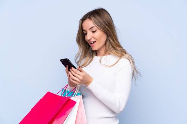 Elinde alışveriş poşetleri olan ve cep telefonuyla bir arkadaşına mesaj yazan izole edilmiş mavi arka planda genç bir kadın.