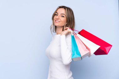 Soyutlanmış mavi arka planı olan genç bir kadın alışveriş torbaları tutuyor ve gülümsüyor.