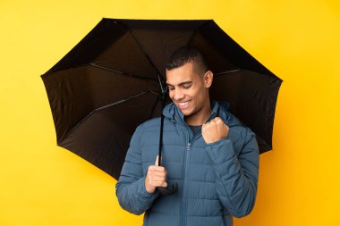 Sarı arka plan üzerinde şemsiyesi olan yakışıklı genç bir adam zaferi kutluyor.