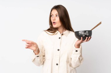 Beyaz arka planda izole edilmiş genç beyaz bir kız elinde çubuklarla bir kase erişte tutarken şaşırmış ve parmağıyla kenarı işaret ediyor.