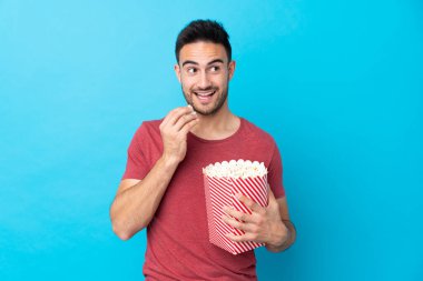 Soyutlanmış mavi arka planda genç yakışıklı bir adam elinde koca bir kova patlamış mısır tutuyor.