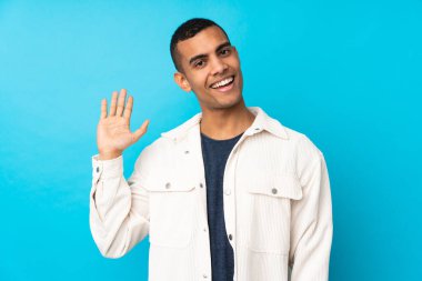 Genç Afro-Amerikalı adam mavi arka planda mutlu bir ifadeyle selamlıyor.