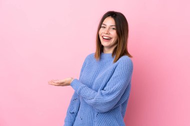 İzole edilmiş pembe arka planda mavi kazaklı genç bir kadın. Davet edilmek için ellerini yana uzatıyor.