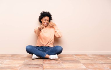 Afrika kökenli Amerikalı bir kadın yerde oturuyor telefon hareketlerini yapıyor ve ön tarafı işaret ediyor.