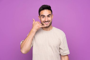 İzole edilmiş mor arka planda genç yakışıklı bir adam telefon jesti yapıyor. Bana geri dönüş işareti ver.