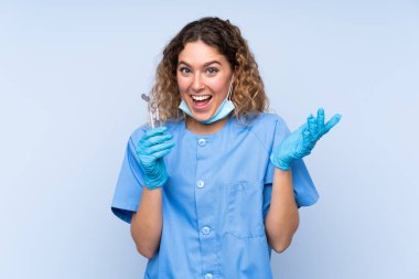 Sarışın kadın diş hekimi mavi arka planda şaşırtıcı bir yüz ifadesi olan aletler tutuyor.