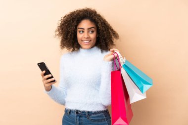 Genç Afrikalı Amerikalı bir kadın bej arka planda izole edilmiş. Elinde alışveriş çantaları ve cep telefonu var.