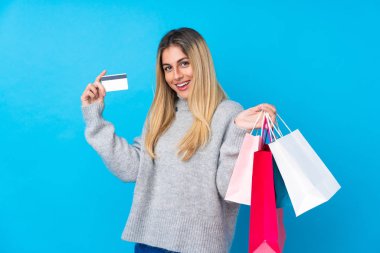 Genç Uruguaylı kadın izole edilmiş mavi arka planda elinde alışveriş çantaları ve kredi kartı tutuyor.
