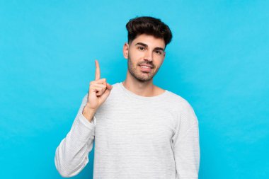 İzole edilmiş mavi arka planı olan genç adam en iyinin işareti olarak parmağını kaldırıyor ve gösteriyor.
