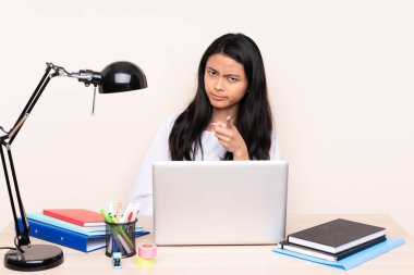 İşyerinde bej arka planda izole edilmiş dizüstü bilgisayarı olan Asyalı kız hayal kırıklığına uğramış ve ön tarafı işaret ediyor.