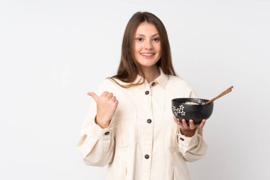 Beyaz arka planda izole edilmiş genç beyaz bir kız elinde çubuklarla elinde bir kase erişte tutarken bir ürünü sunmak için kenarı işaret ediyor.