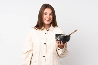 Beyaz arka planda, elinde yemek çubuklarıyla elinde bir kase şehriye tutarken, şaşırmış ve şok olmuş bir yüz ifadesi olan beyaz bir kız.