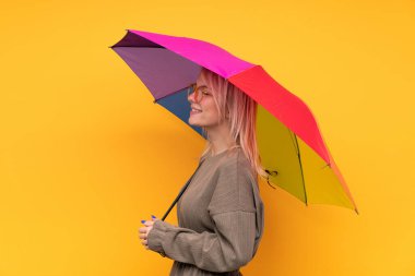 Sarışın genç bir kadın şemsiyesini izole edilmiş sarı duvarın üzerinde tutuyor ve yan tarafa bakıyor.